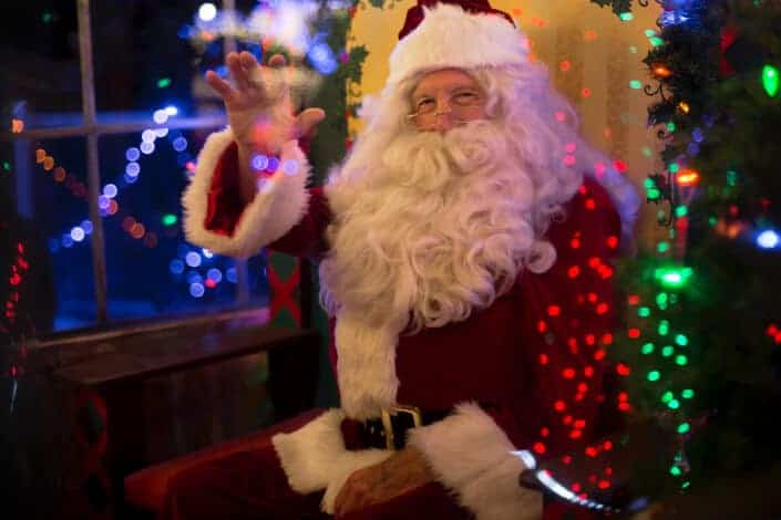 Waving Santa beside a Christmas tree, pitching corny jokes.